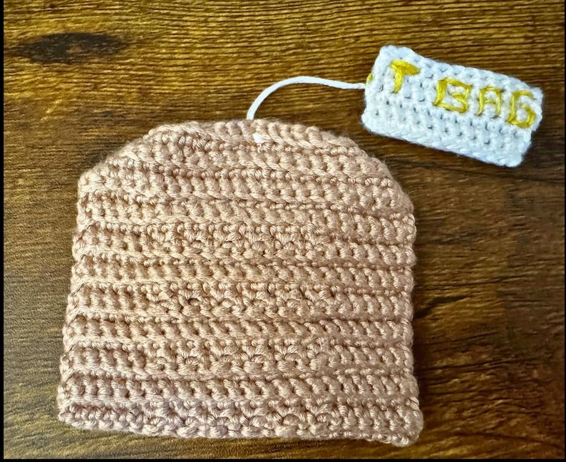 May include: A light brown crocheted tea bag cosy, with a matching crocheted tag that reads "TEA BAG" in yellow thread. The cosy and tag are connected by a white string.