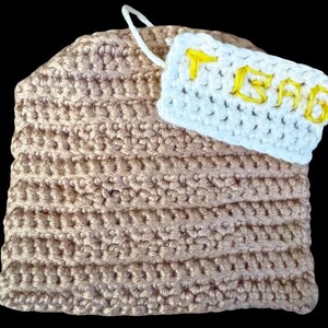 May include: A light brown crochet tea bag with a white tag. The tag has the word "TEA" embroidered in yellow. A white string is attached to the tag. The tea bag is handmade.