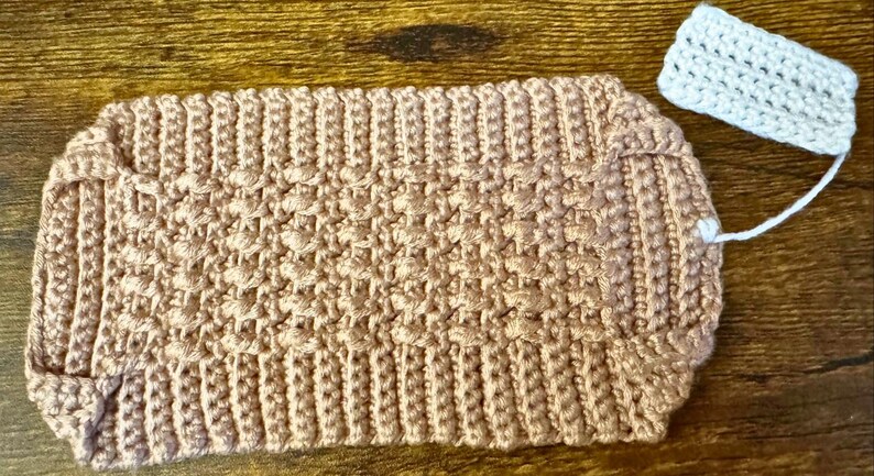 May include: A handmade, light brown crochet pouch with a textured stitch pattern. A small, rectangular, white crochet tag is attached with a white string. The pouch is likely used for small items.