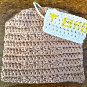 May include: A light brown crocheted tea cosy. A white crocheted tag is attached, displaying the word "TEA" in yellow yarn. The cosy is rectangular with a rounded top and a hanging loop.