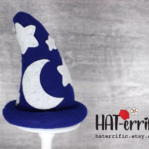 May include: A blue felt wizard hat with a white crescent moon and stars. The hat is on a white stand. The text "HAT-errific" is in the lower right corner of the image.