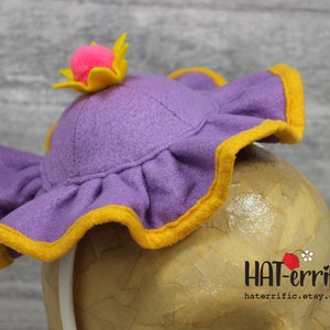 May include: Purple felt hat with a yellow ruffle trim and a pink pom-pom on top. The hat is on a brown mannequin head.