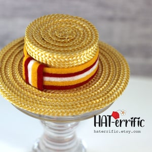 May include: A straw boater hat with a yellow, red, and white striped band. The hat has a round crown and a wide brim. The hat is displayed on a clear glass stand. The text "HAT-errific" and the Etsy shop URL are visible.