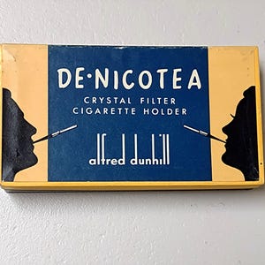 May include: A yellow and blue box with the text "DE-NICOTEA CRYSTAL FILTER CIGARETTE HOLDER alfred dunhill" and a black silhouette of a person smoking a cigarette on each side.