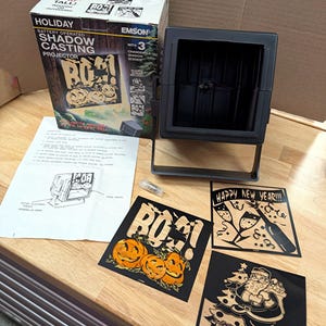 May include: A black battery-operated shadow casting projector with its original box, an instruction manual, and three interchangeable shadow scene slides featuring a pumpkin design with the text BOO!, a Happy New Year design, and a Santa Claus design.