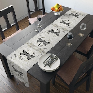 May include: A rectangular table runner with a dragonfly pattern. The runner is white with a distressed, textured appearance and black dragonfly illustrations. The table is dark gray, set with plates, silverware, wine glasses, and a bowl of fruit.