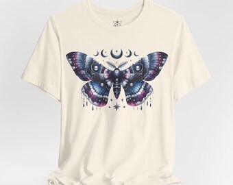 Puede incluir: Camiseta color crema con un diseño grande de polilla. La polilla tiene alas azules, moradas y blancas con un diseño de luna creciente y estrellas encima. El diseño está centrado en la parte delantera de la camiseta.