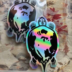 May include: Holographic stickers featuring a turtle design with a world map on its shell. The black outline of the turtle and the map contrasts with the iridescent background. The stickers are in a clear plastic bag, with a few stickers visible.