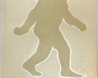 May include: A beige Bigfoot silhouette walking to the right, with the word "BELIEVE" in a matching beige font below. The image has a textured, slightly speckled appearance.