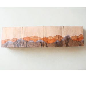 Puede incluir: Una pieza de arte mural de madera rectangular y larga con un diseño de paisaje. La obra de arte representa una cordillera en tonos de naranja, gris y marrón sobre un fondo claro. La pieza está montada en una pared blanca.