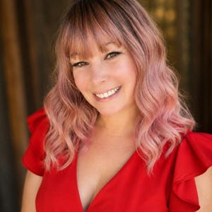 May include: A person with shoulder-length wavy hair dyed in a soft pastel pink hue, featuring straight-cut bangs that frame the forehead. The individual is wearing a bright red top with short, ruffled sleeves and a deep V-neckline, smiling directly.