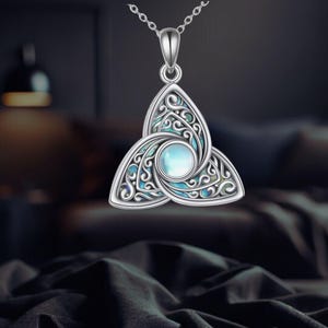 Puede incluir: Collar con colgante plateado con diseño de nudo celta. El colgante presenta una piedra central azul-verdosa y un intrincado trabajo de volutas. El collar se muestra sobre un fondo oscuro.