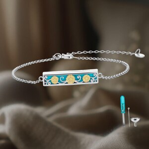 May include: Silver bracelet featuring a rectangular pendant with a turquoise background and three gold-colored sunflowers. The bracelet has a delicate chain and a heart-shaped clasp. Includes a small blue screwdriver and funnel.
