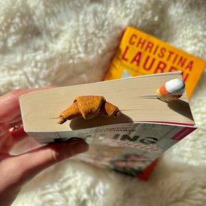May include: A handmade croissant bookmark and a candy corn bookmark are inserted into a book. The book has a white cover with the title "Falling in Love" and the author's name. A second book with a yellow cover is visible in the background.