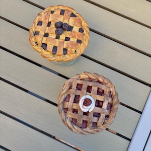 Peut inclure: Deux petites tartes avec des croûtes en treillis. L'une a une garniture aux myrtilles et trois myrtilles sur le dessus. L'autre a une garniture aux cerises et un tourbillon de glaçage blanc. Les tartes ont une croûte brun doré.