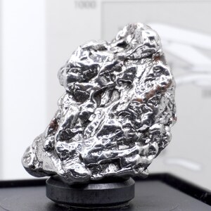 May include: A metallic, silver-colored iron meteorite specimen with a highly irregular, textured surface featuring deep ridges, pits, and a reflective, polished appearance. The object is mounted on a small, circular black base for display purposes.