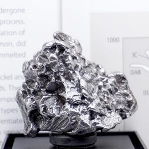 May include: A metallic, silver-toned iron meteorite specimen with a highly irregular, pitted, and textured surface. The object is mounted on a small, circular black base, positioned in front of a white informational card with printed text and a diagram.