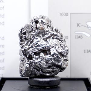 May include: A metallic, silver-coloured iron meteorite specimen with a highly irregular, pitted, and textured surface. The object is mounted on a small, circular black base, displaying a reflective, lustrous finish across its complex, jagged form.