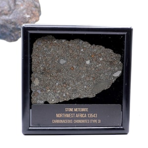 May include: A slice of Northwest Africa 13543 carbonaceous chondrite stone meteorite displayed in a square black frame. The specimen features a textured surface with numerous small, multicoloured inclusions. A label reads: STONE METEORITE NORTHWEST AFRICA 13543 CARBONACEOUS CHONDRITES (TYPE 3).