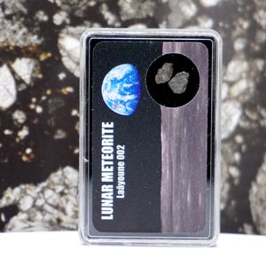 May include: A clear plastic case containing a lunar meteorite specimen. The card inside features a blue and white image of Earth, the words "LUNAR METEORITE Laâyoune 002", and a small circular window revealing two gray meteorite fragments.