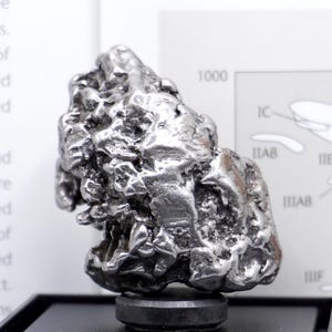 May include: A metallic, silver-coloured iron meteorite specimen with a highly irregular, pitted, and textured surface. The object is mounted on a small, circular black base, positioned in front of a white informational card with printed text and diagrams.