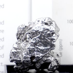 May include: A metallic, silver-colored iron meteorite specimen with a highly irregular, pitted, and textured surface. The object is displayed in front of a white informational card featuring printed text and a chart labeled Ge (ppm).