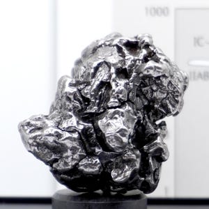 May include: A metallic iron meteorite specimen with a dark, irregular, and pitted surface texture. The object features a complex, craggy form with deep crevices and raised ridges, displaying a reflective, gunmetal grey finish throughout its structure.