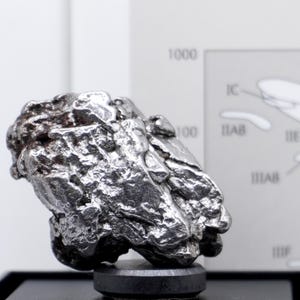 May include: A metallic, silver-coloured iron meteorite specimen with a highly irregular, pitted, and textured surface. The object is mounted on a small, circular black base, positioned in front of a printed diagram featuring text and labels.