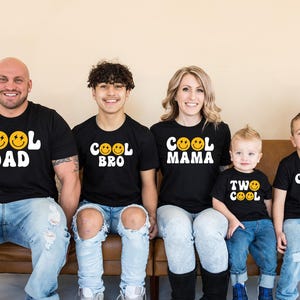 May include: A family of five wearing matching black t-shirts with a yellow smiley face and the words "Cool Dad", "Cool Bro", "Cool Mama", "Two Cool", and "Cool Bro" printed on them. The family is sitting on a brown couch.