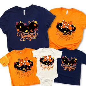 May include: A collection of navy blue, orange, and white t-shirts and a onesie. Each garment features a black Mickey Mouse-shaped design with an orange bow, fall leaves, and the word "Thankful" in a script font.