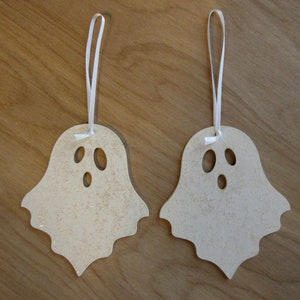 May include: Two wooden ghost ornaments with a light beige color and glitter accents. Each ghost has three oval cut-out eyes and a mouth. White ribbon is attached for hanging, perfect for Halloween decorations.