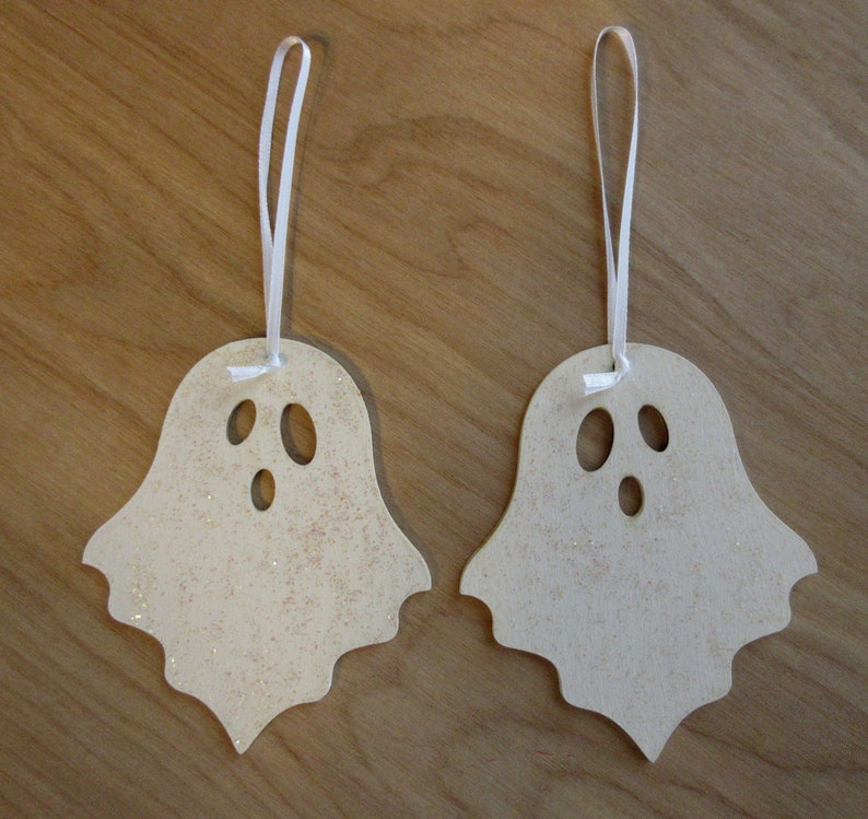 May include: Two wooden ghost ornaments with a light beige color and glitter accents. Each ghost has three oval cut-out eyes and a mouth. White ribbon is attached for hanging, perfect for Halloween decorations.