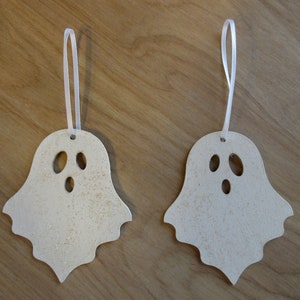 May include: Two wooden ghost ornaments with three eye holes and a mouth cutout. Each ghost has a white ribbon for hanging. The ghosts are light beige with gold glitter accents, perfect for Halloween decorations.