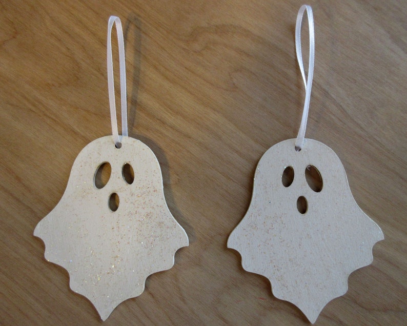 May include: Two ghost-shaped ornaments with a light beige color and glitter accents. Each has a white ribbon for hanging and three oval cutouts for eyes and mouth. Perfect for Halloween decorations.