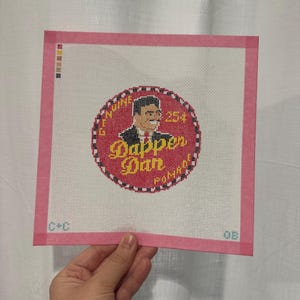 May include: A square needlepoint canvas featuring a circular design with a pink background. The center displays a man in a suit with the text Dapper Dan in yellow, along with the words Genuine, Pomade, and the number 254. The canvas has a pink border.