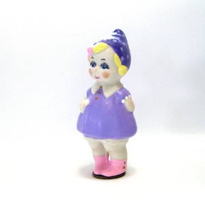 May include: A vintage ceramic figurine of a young girl. The figurine is wearing a purple dress, a blue hat with white polka dots, and pink boots. The girl has rosy cheeks, blue eyes, and yellow hair. The figurine is standing on a white surface.