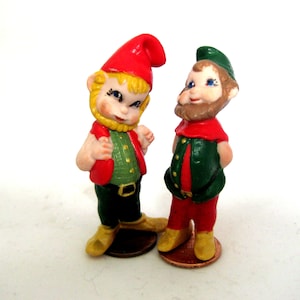 May include: Two small ceramic figurines of gnomes. The gnome on the left is wearing a red hat, a green vest, and green pants. The gnome on the right is wearing a green hat, a green vest, and red pants.