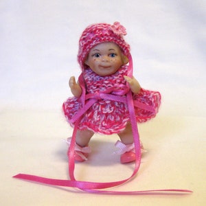 May include: A small porcelain doll wearing a pink and white knitted dress and hat with a pink ribbon. The doll is wearing pink shoes and has a flower on its hat.