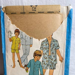 Puede incluir: Paquete de patrones de pijamas vintage con ilustraciones de niños en varios estilos de pijamas. Los diseños incluyen colores lisos, rayas y telas estampadas. El paquete está en una funda de cartón azul y blanco.