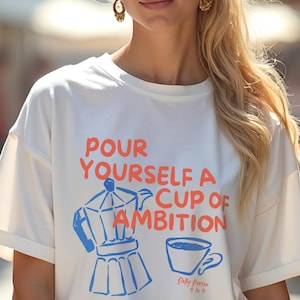 Könnte beinhalten: Eine Frau trägt ein weißes T-Shirt mit dem Text "Pour Yourself a Cup of Ambition" in Orange und Blau. Das Shirt zeigt eine Kaffeekanne und eine Kaffeetasse. Die Frau hat blondes Haar und trägt goldene Ohrringe.