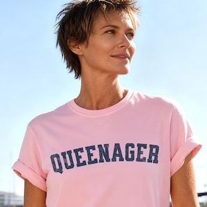 Puede incluir: Una camiseta rosa claro con la palabra "QUEENAGER" impresa en letras de bloque azul marino. La camiseta tiene las mangas enrolladas. El fondo muestra barcos en el agua y un cielo azul claro.
