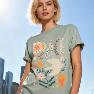 May include: Sage green t-shirt with rolled sleeves featuring the word "PEACE" in a whimsical font, along with a dove and floral graphics. The shirt is worn outdoors with a city and water background.