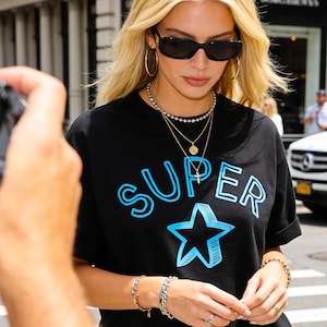 Può includere: T-shirt nera con la scritta "SUPER" in azzurro chiaro, bordata di blu più scuro, sopra una stella blu. La persona indossa occhiali da sole, orecchini a cerchio dorati e diverse collane e bracciali. Un'auto bianca è sullo sfondo.