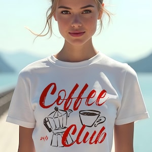 May include: White t-shirt with the words "Coffee Club" in red script, along with a red illustration of a coffee pot and a coffee cup. The shirt is a crew neck style.