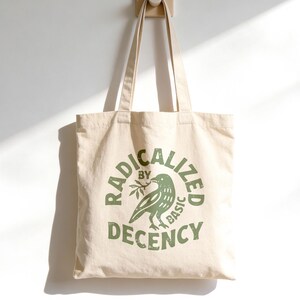 Pode incluir: Uma sacola tote creme com as palavras "RADICALIZED by BASIC DECENCY" em verde, junto com uma ilustração de um pássaro segurando um ramo de oliveira. A sacola está pendurada em um gancho de madeira contra uma parede branca.
