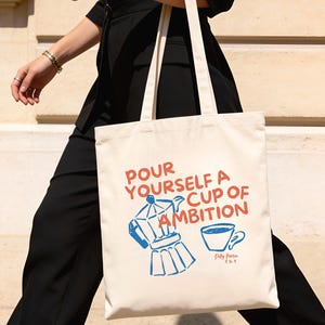 May include: Cream-colored tote bag with the text "Pour Yourself a Cup of Ambition" in red and blue. The bag features a blue coffee maker and a coffee cup graphic. The bag is carried by a person walking outside.