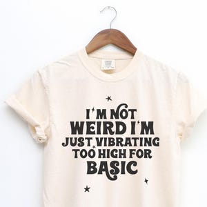 May include: Cream-colored t-shirt with the phrase "I'm not weird I'm just vibrating too high for basic" in black lettering. The shirt is hanging on a wooden hanger against a white background. Small star graphics are also present.