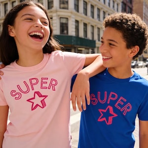 Puede incluir: Dos niños con camisetas a juego con la palabra "SUPER" y una estrella. Una camiseta es rosa, la otra azul. Están en una calle de la ciudad, sonriendo y riendo.