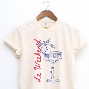 Ivory Beige T-shirt with the text Le Weekend and a Cherry Coquette Cocktail graphic.

Because what’s a good time without a little flair?

Perfect for brunch dates, rooftop soirées, or just channeling that effortlessly flirty energy. French twist, retro feel, full-on main character mood.
Weekend Shirt, Le Weekend, French T-Shirt, Weekending, Vintage Aesthetic, Coquette Bow Cherry, Cocktail Tee, y2k T-shirt, weekend vibes tee.