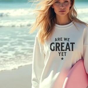 Op de afbeelding: Een vrouw draagt een wit sweatshirt met de tekst "ARE WE GREAT YET" in zwarte letters en een ster. Ze houdt een roze surfboard vast op een strand.